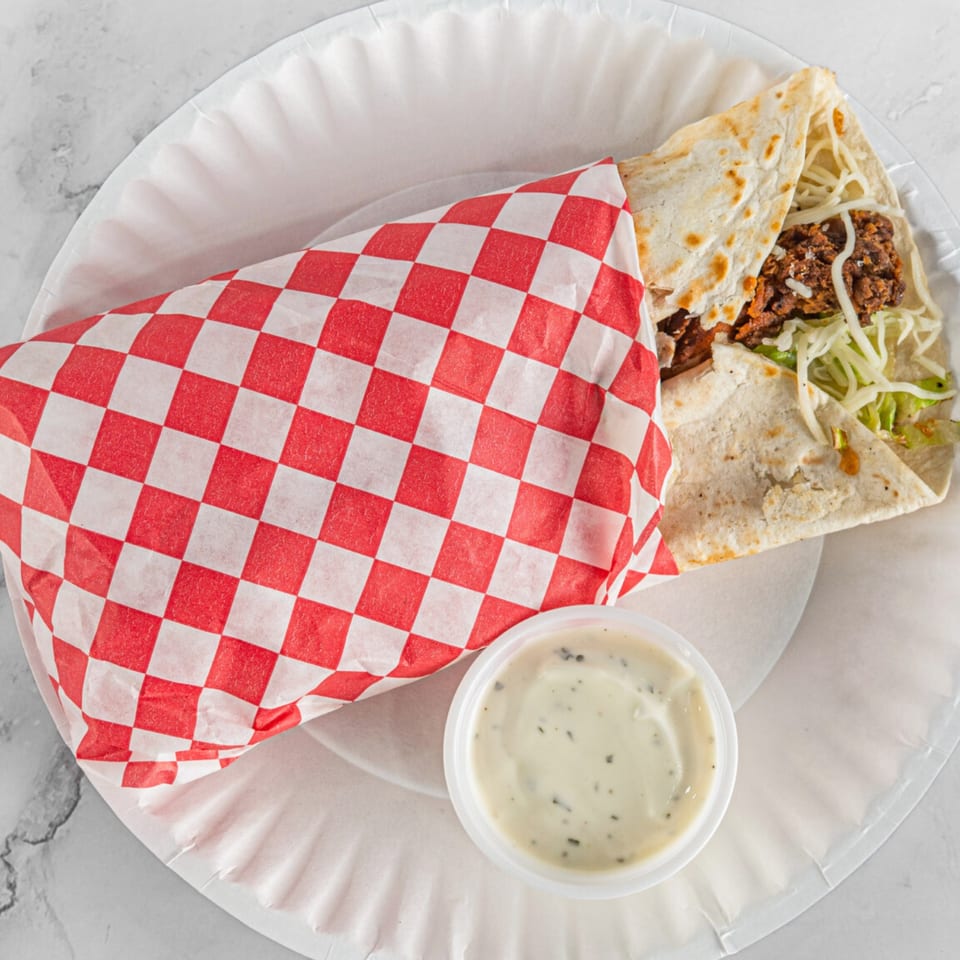 A Hot Chicken and Breakfast burrito, partially wrapped in red and white checkered paper, with a side of sauce.